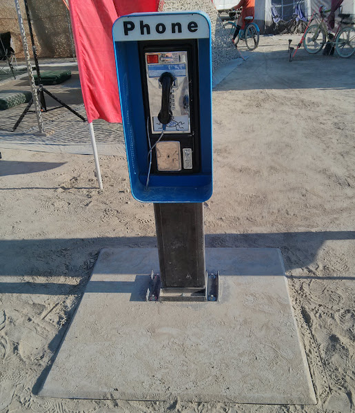 The Playa Phone booth on the playa: a blue pay phone on a black post, with a PHONE sign on top, in front of camp shade and bikes.
